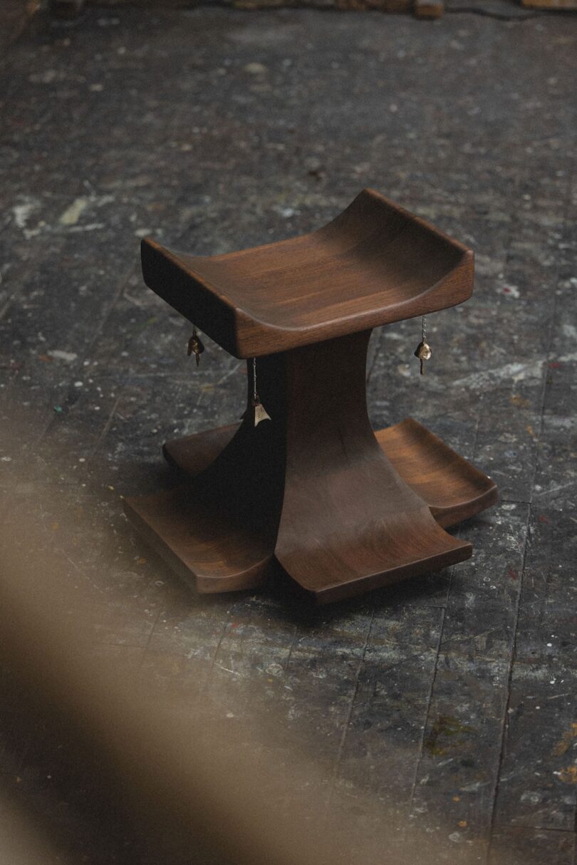 Un taburete escultural de madera oscura con asiento y base curvos se asienta sobre un piso desgastado y salpicado de pintura; baratijas cuelgan de los bordes.
