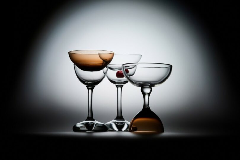 Tres vasos cupé están dispuestos uno al lado del otro; dos están derechos y uno invertido. Un vaso contiene cerezas rojas, los otros dos tienen un sutil color ámbar.