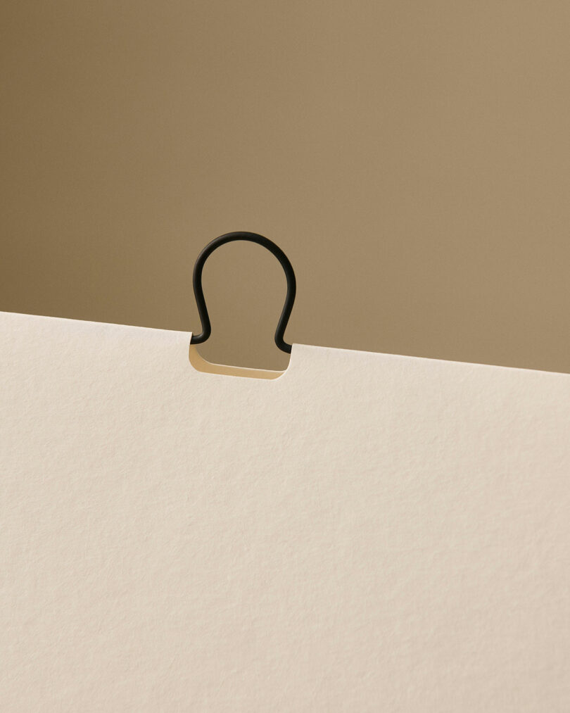 Una carpeta negra fijada con alfileres en el borde superior de un trozo de papel blanquecino sobre un fondo beige.