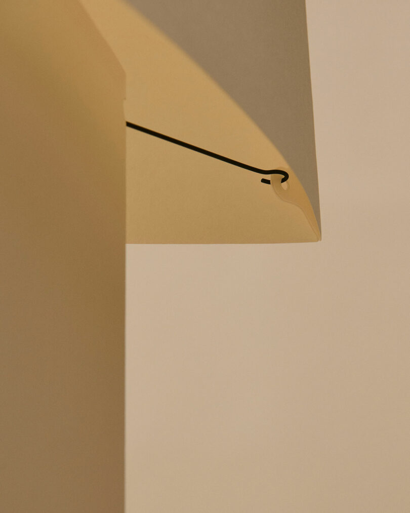 Primer plano de una pantalla de lámpara beige con un fino cable de soporte metálico visible unido a su borde interior, sobre un fondo neutro.