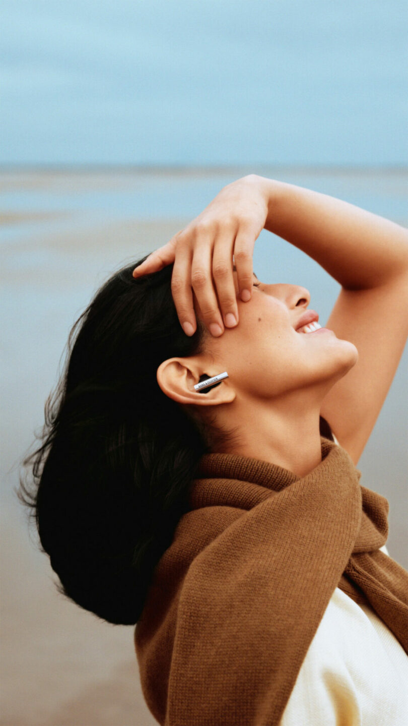 Un hombre que lleva un pañuelo marrón está al aire libre con la cabeza inclinada hacia atrás y una mano en la frente sonriendo. El fondo presenta un paisaje tranquilo y abierto.