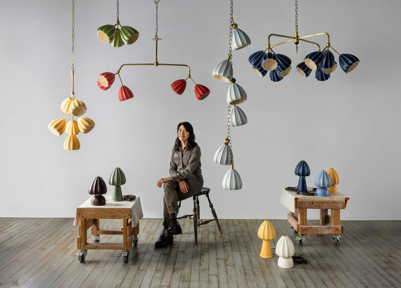Hay una mujer sentada en un taburete en la habitación, y hay candelabros modernos con forma de flor y luces de varios colores colgando del techo sobre dos mesas.