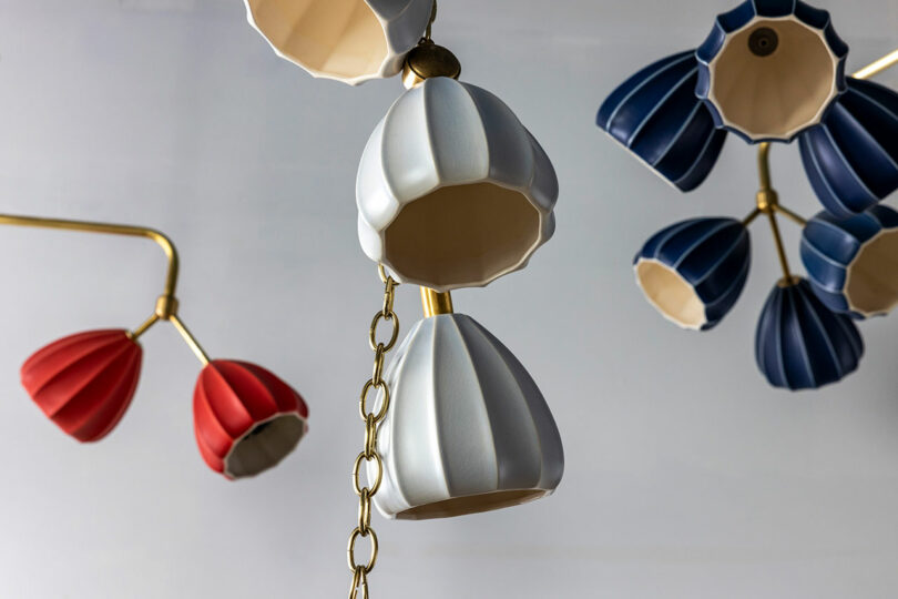 Lámpara de araña moderna con pantallas de cerámica en forma de abanico blancas, azules y rojas suspendidas de accesorios de metal dorado sobre un fondo claro.