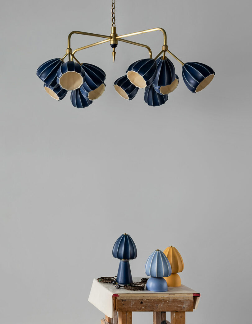 Una lámpara de araña de latón con una pantalla azul en forma de pétalo cuelga sobre la mesa, que está rematada con lámparas de mesa azules y amarillas que combinan con las paredes grises lisas.