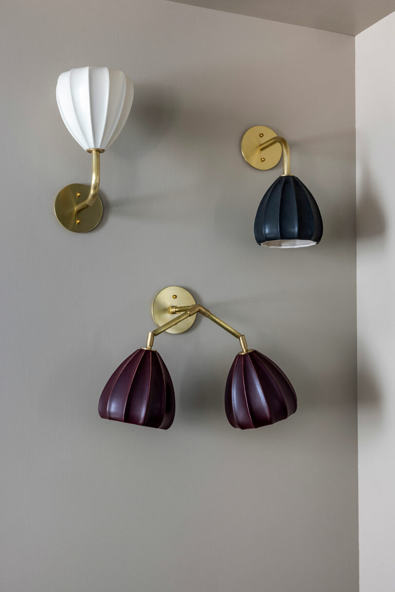 Tres lámparas de pared con pantallas plisadas en blanco, negro y burdeos están montadas en paredes de color gris claro con herrajes de latón.