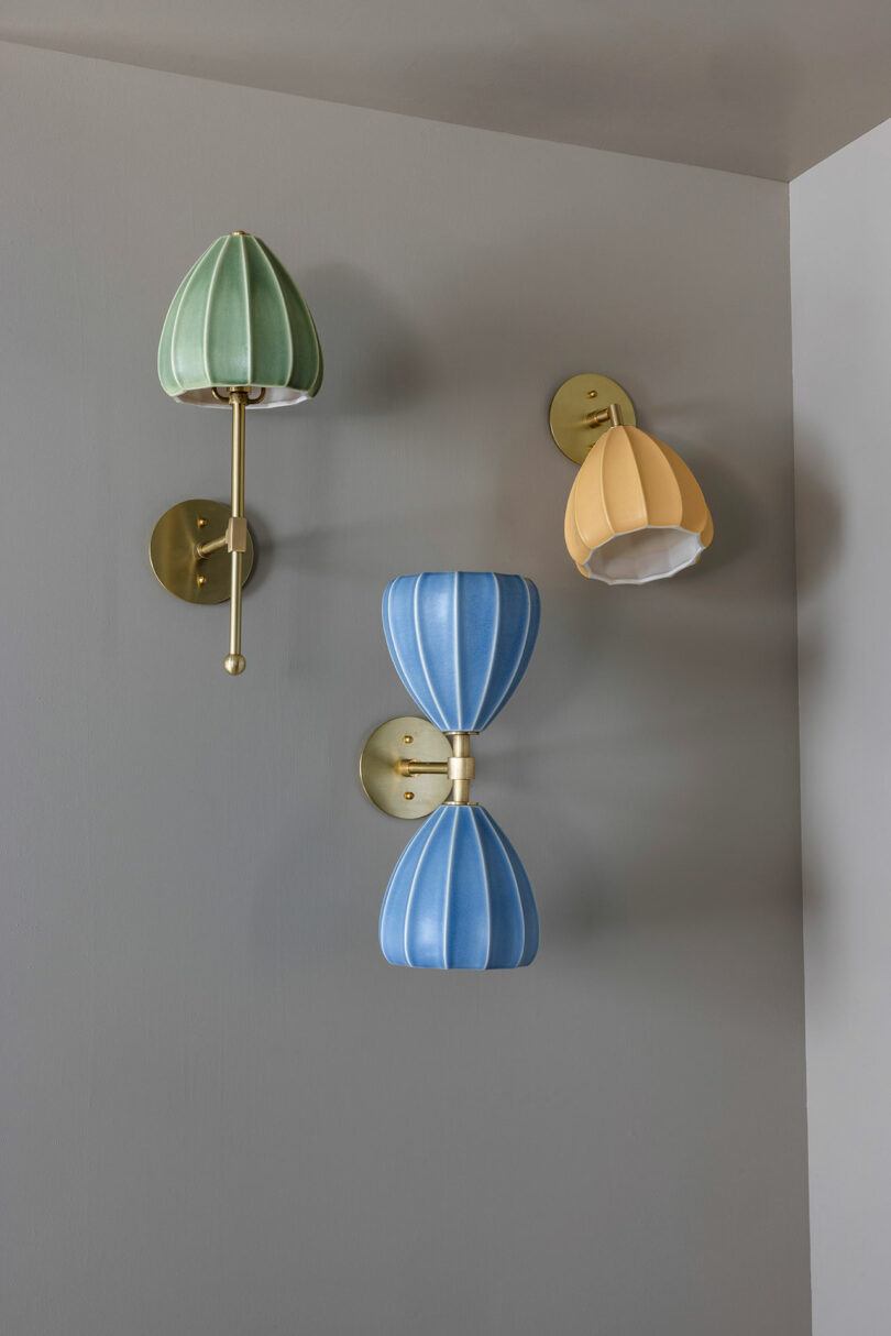 Tres lámparas de pared con accesorios dorados, con tonos verdes, amarillos y azules, se alinean en la pared gris claro.