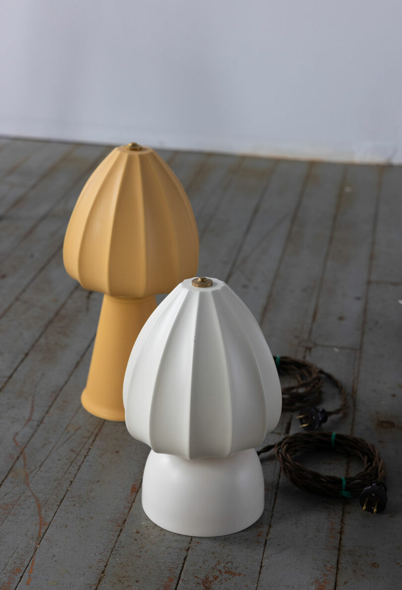 Dos lámparas con forma de hongo, una amarilla y otra blanca, se encuentran en el suelo de madera junto a cables de alimentación enrollados.
