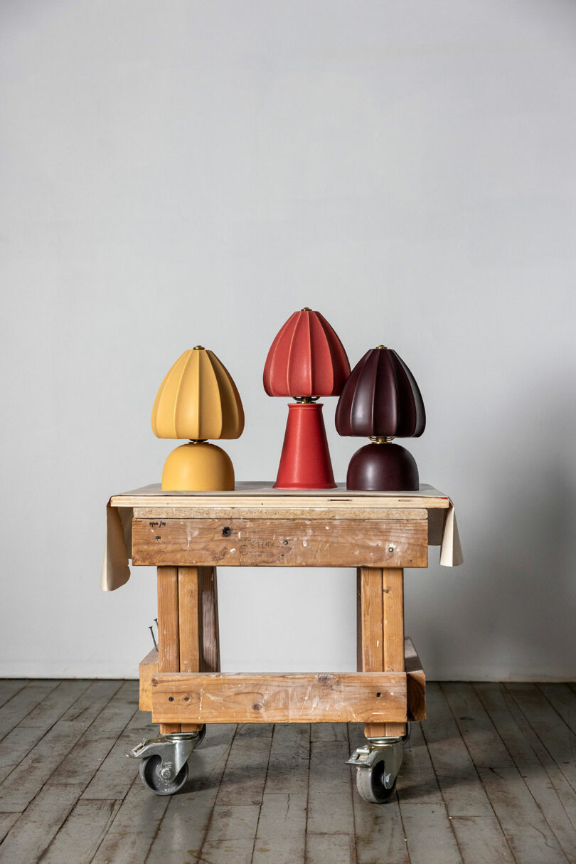 Tres lámparas de mesa en forma de cúpula de color amarillo, rojo y marrón oscuro se exhiben en un carrito de madera contra una pared gris claro.