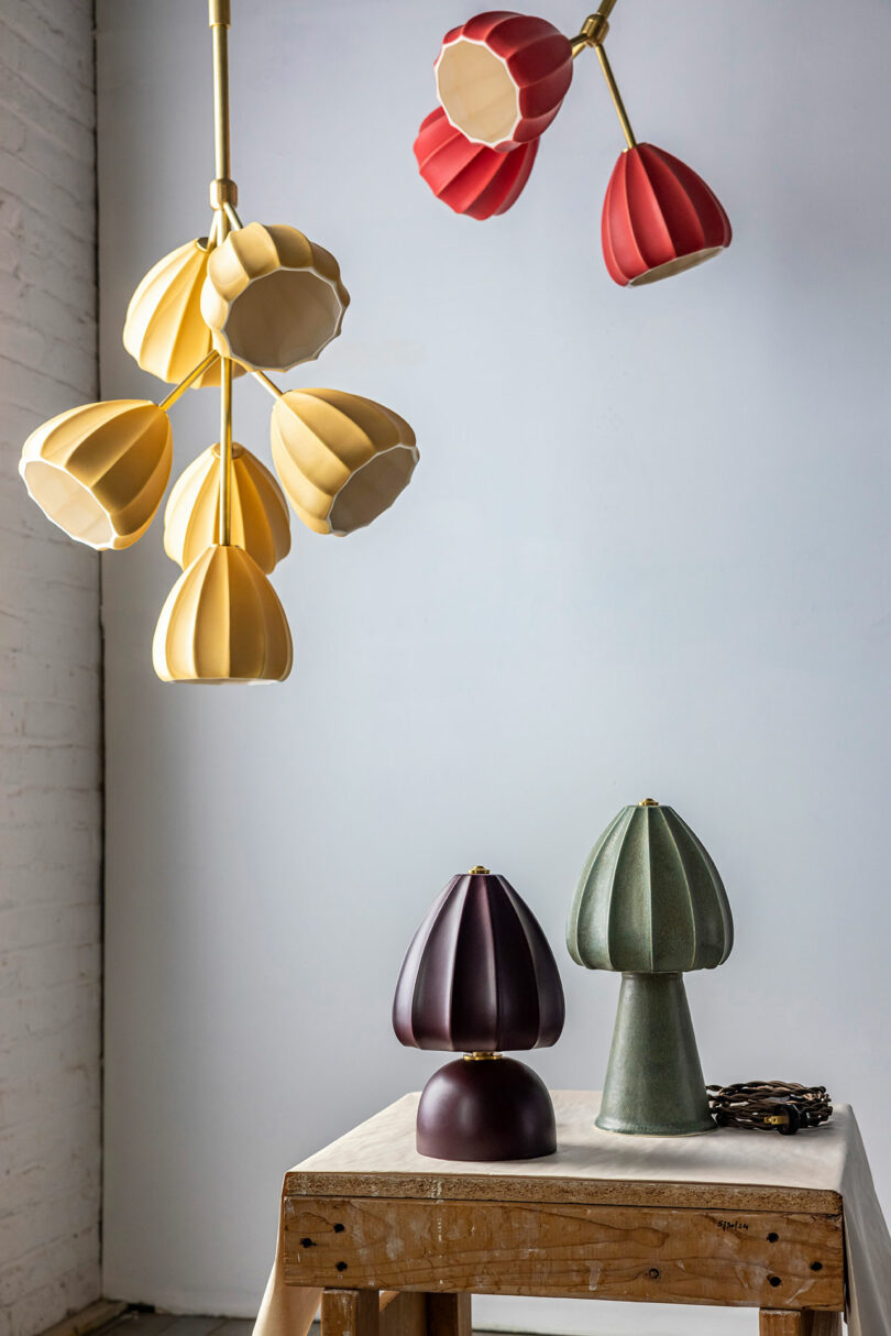 Candelabros amarillos y rojos cuelgan sobre mesas con lámparas verdes y burdeos en forma de hongo, sobre un fondo de paredes y ladrillos blancos.