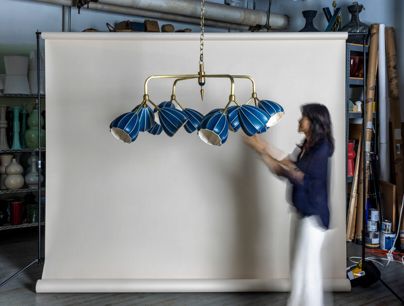 Una mujer ajusta una lámpara de araña de latón con ocho tonos azules frente a un fondo beige en un ambiente de estudio.