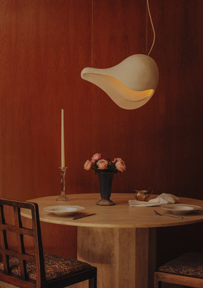 Mesa redonda para dos con platos, cubiertos, un jarrón de flores rosas, un candelabro y una singular lámpara de araña blanca en la pared de madera.