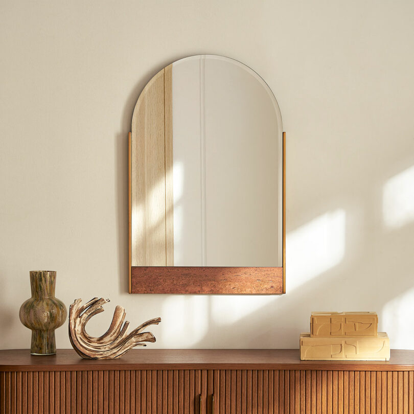 A wooden sideboard with a green vase, sculptural wooden object, stacked books, and an arched wall mirror above—perfect for displaying Valentine's Day gifts—set against a beige wall.