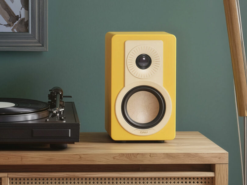 A yellow DALI bookshelf speaker sits on a wooden table next to a vinyl record player, against a green wall.