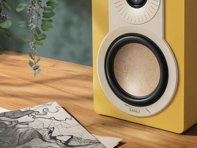 A yellow DALI speaker rests on a wooden surface beside a black and white abstract drawing, with vibrant green leaves hanging on the left.