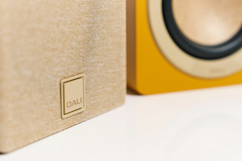 Close-up of a beige DALI speaker with its logo, alongside part of a yellow speaker featuring a visible woofer in the background, all placed on a white surface.