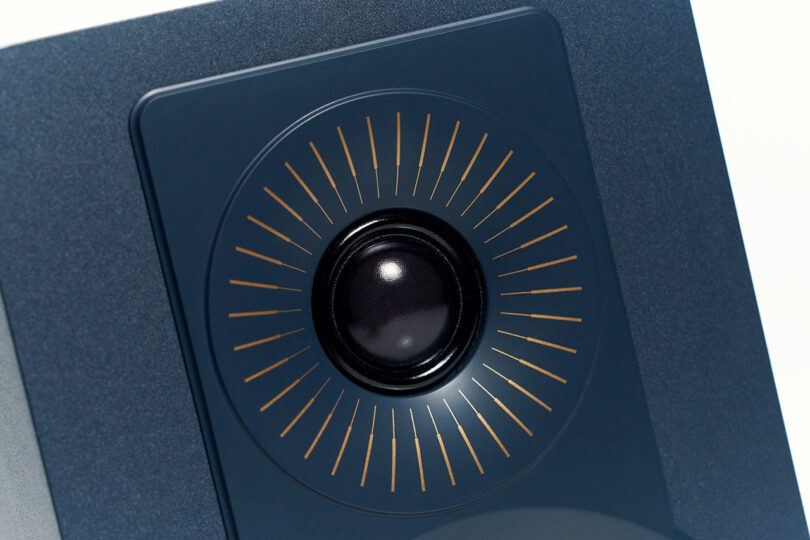 Close-up of a DALI speaker with a dark circular driver, surrounded by a square panel and radiating gold lines on a dark background.