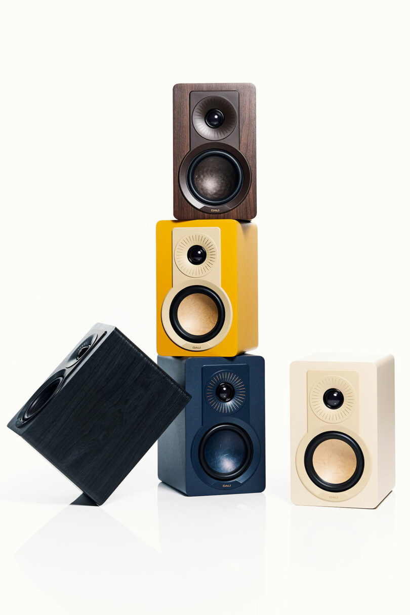 Four modern DALI speakers in brown, yellow, blue, and cream are stacked vertically, with a black speaker angled and leaning against the stack.