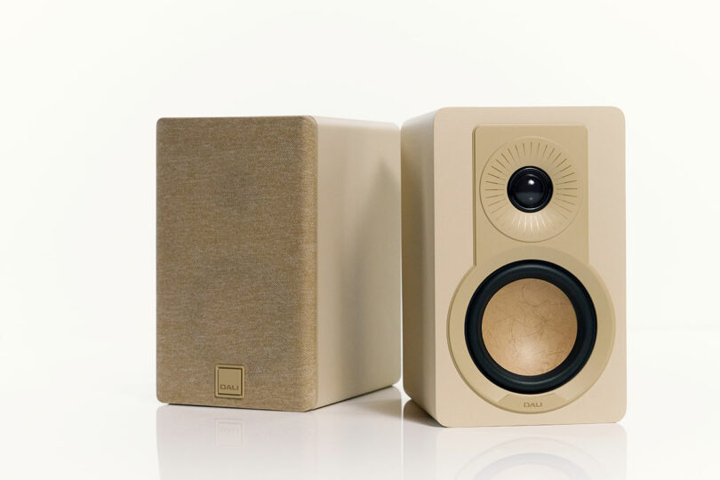 Two beige-colored DALI bookshelf speakers are shown; one with the front facing forward displaying drivers, the other with the back facing out covered by a fabric grille.