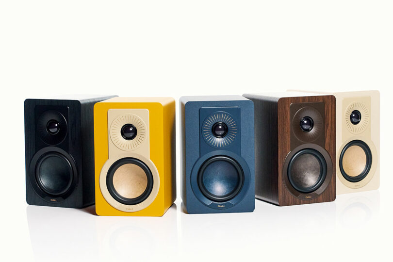 Four DALI bookshelf speakers in black, yellow, blue, and wood finish are arranged side by side on a white background.