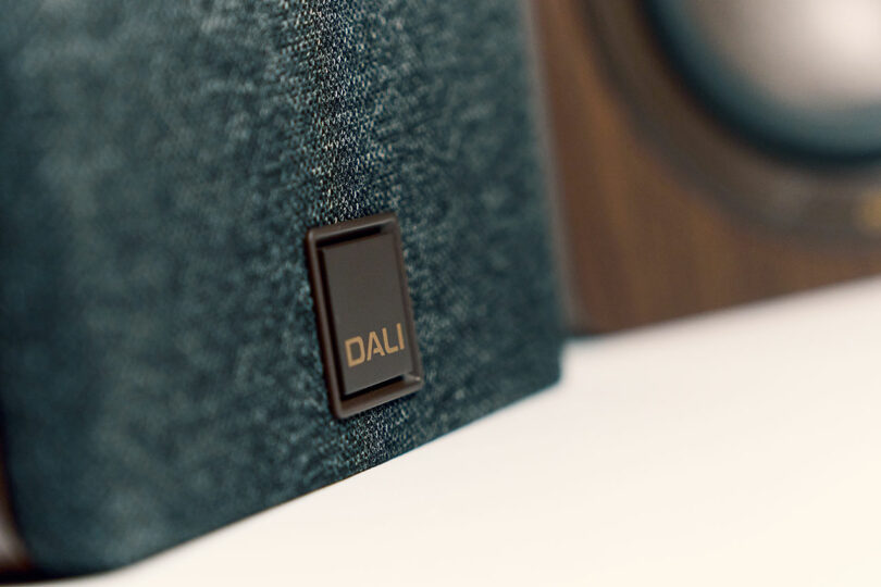 Close-up of a DALI speaker with a fabric grill and the DALI logo in focus; another wood-finished speaker is softly blurred in the background.