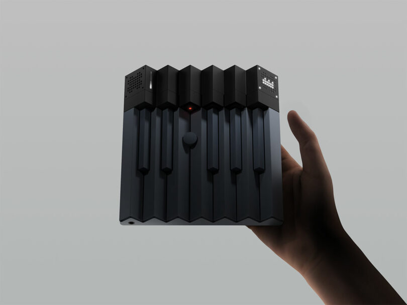 A hand holds a compact, black electronic device with piano-like keys and small LED screens on the top edge against a plain gray background.