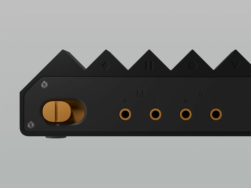 A close-up of a black electronic device with geometric buttons, four gold audio jacks labeled "+", "M", "-", and "A", and a gold rotary knob on the left side.