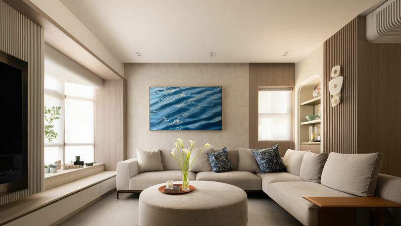 Modern living room with a beige sectional sofa, round ottoman, decorative pillows, a wall-mounted abstract blue art piece, and natural light from large windows.