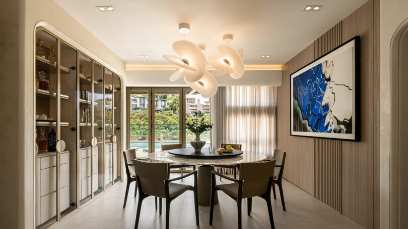 A modern dining room with a round table, six chairs, a sculptural chandelier, large windows, glass-front cabinets, and abstract art on the wall.