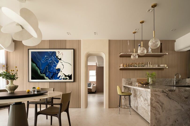 Modern kitchen and dining area with marble countertops, wooden walls, abstract wall art, pendant lights, open shelves, and contemporary furnishings.