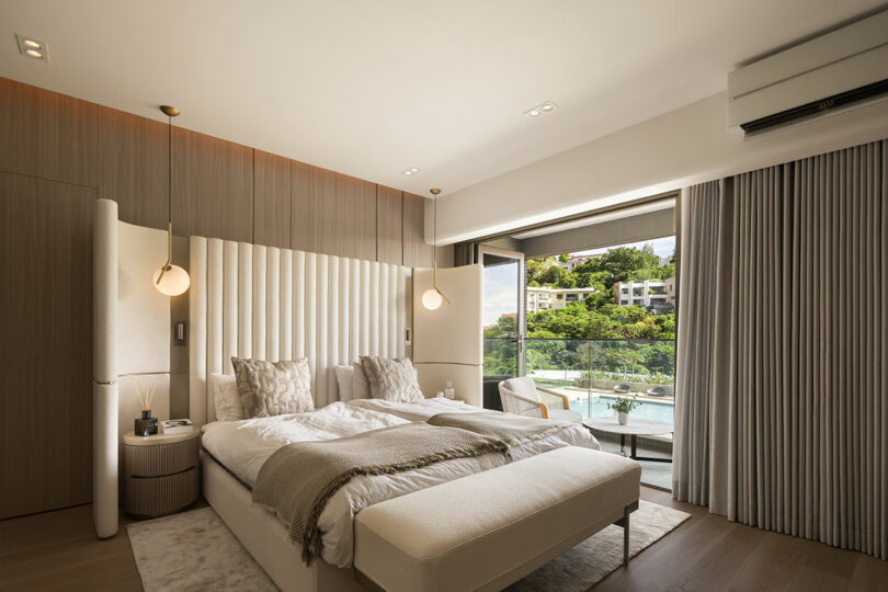 Modern bedroom with neutral tones, twin beds, upholstered headboard, pendant lights, large window, balcony, and view of greenery and buildings outside.