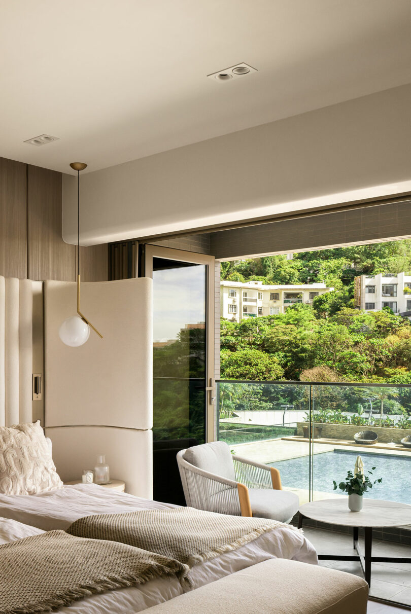 A modern bedroom with neutral decor, two beds, a chair, a round table, and large glass doors opening to a balcony with a view of greenery and nearby buildings.