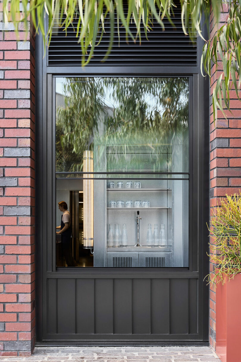 View through a large window shows shelves with glassware and water bottles; a person stands in the background, partially visible. Brick and metal window frame, greenery above.