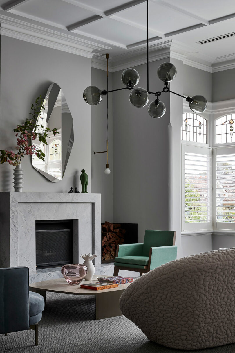 Salón moderno con chimenea de mármol, espejo geométrico, lámpara de araña moderna, sillón verde, sofá texturizado, mesa baja y persianas.