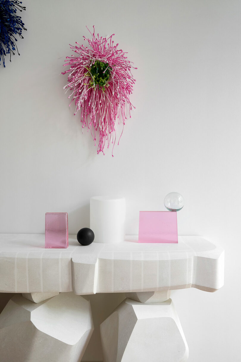 Una mesa geométrica blanca con bloques de vidrio rosa, una esfera negra, una esfera transparente, un cilindro blanco y una escultura floral abstracta rosa montada en la pared.