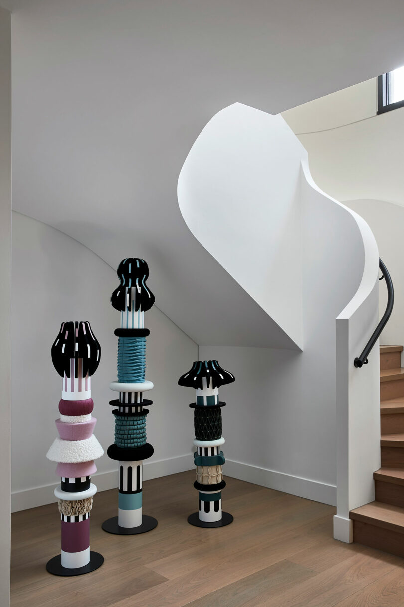 Tres coloridas columnas escultóricas abstractas se encuentran debajo de una moderna escalera blanca con escalones de madera y pasamanos curvos, un interior minimalista.