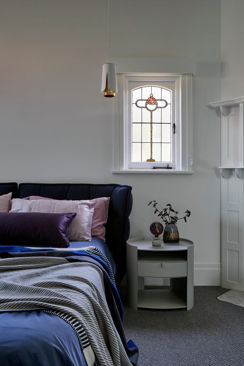 En el dormitorio suavemente iluminado, debajo de una vidriera decorativa, hay una cuidada cama cubierta con sábanas de color violeta y azul, con una pequeña mesita de noche redonda al lado.