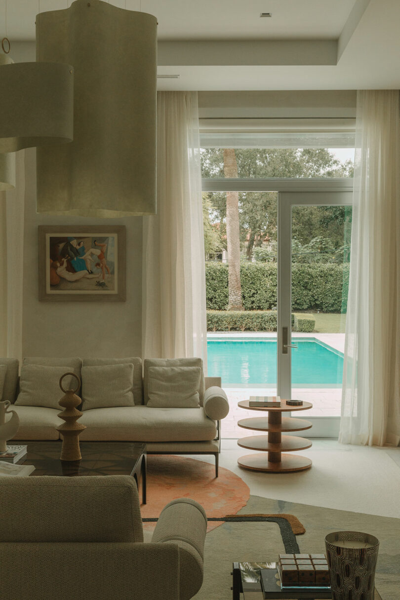 A modern living room with neutral-toned furniture, abstract decor, large windows, and sheer curtains overlooking a backyard with a swimming pool and greenery.