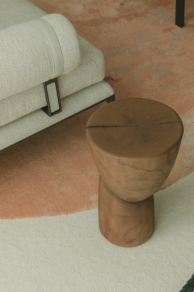 A round wooden stool sits on a light beige and peach rug beside a textured white sofa with a white leather strap detail.