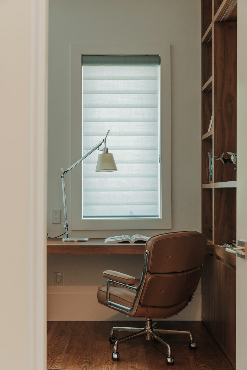 A modern home office with a brown swivel chair, wooden desk, desk lamp, open book, built-in shelves, and a window with a closed blind.
