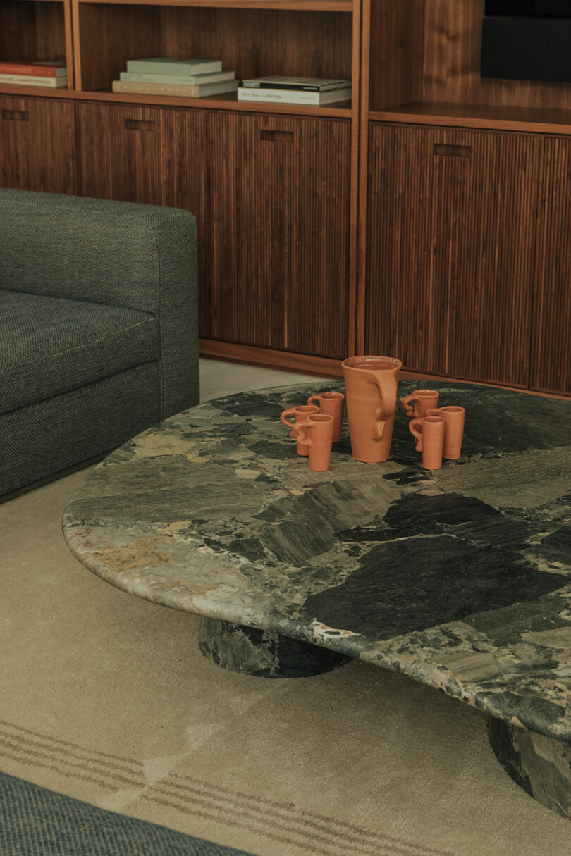 A stone coffee table with five terra cotta mugs and a pitcher sits on a beige rug, next to a gray sofa and a wooden cabinet with books.
