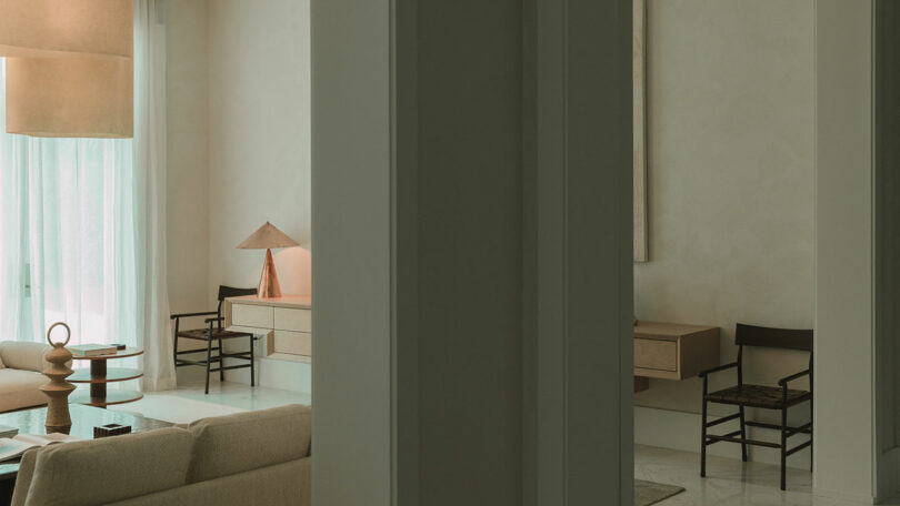 A modern living room with neutral tones, featuring a beige sofa, wooden chairs, a round coffee table, and a desk with a lamp, viewed through an open doorway.