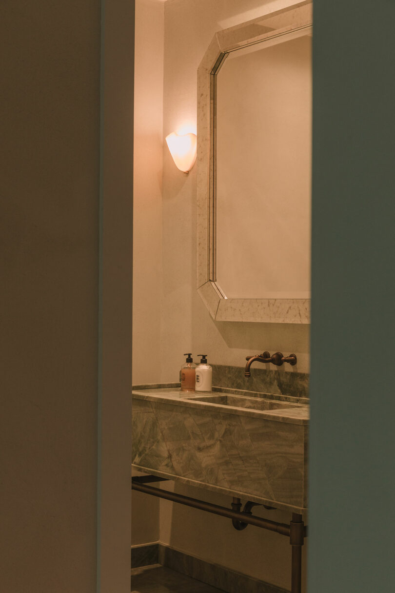 A bathroom with a marble sink, two soap dispensers, a wall-mounted faucet, a large mirror, and a wall sconce light, seen through a doorway.