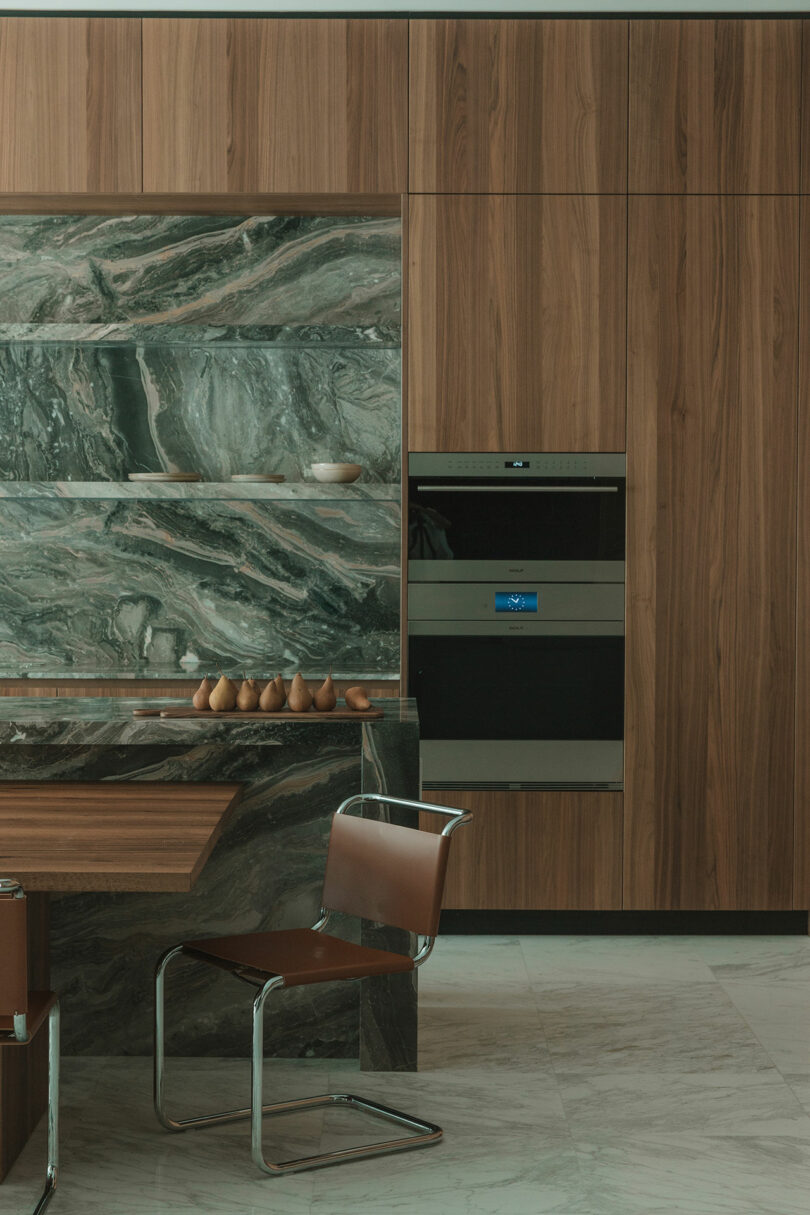 Modern kitchen with wood-paneled cabinets, green marble backsplash and countertop, built-in oven, brown leather chair, and decorative items on a shelf.