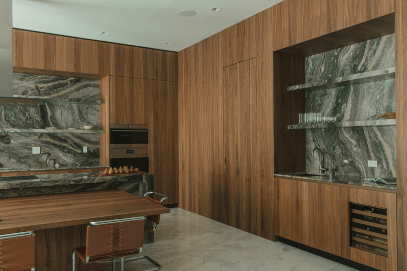 Modern kitchen with wood cabinetry, marble backsplash and countertops, built-in oven, open shelves with glasses, and seating area with brown chairs on a marble floor.