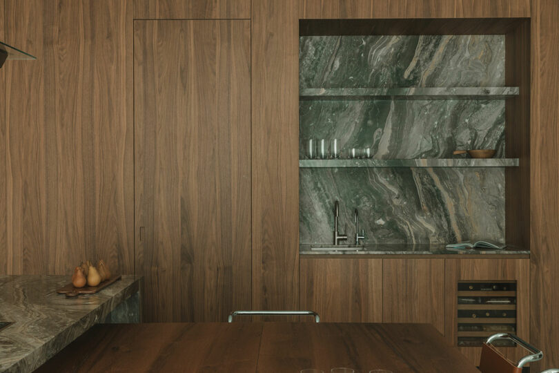 A modern kitchen with wood panel walls, marble countertops and backsplash, open shelves with glasses, a small sink, and a built-in wine fridge.