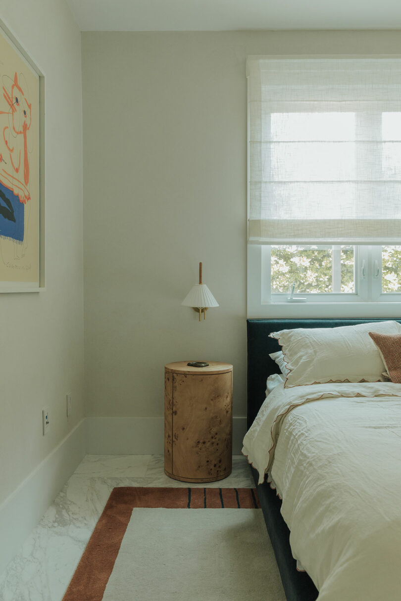 A neatly made bed next to a wooden nightstand with a small lamp, under a window with sheer curtains; modern art hangs on the adjacent wall.