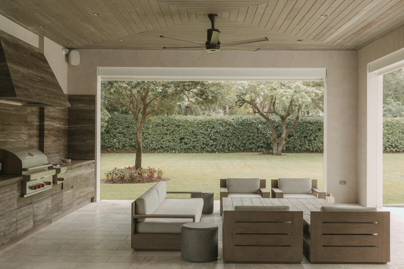 Modern covered patio with built-in grill, ceiling fan, and wooden outdoor furniture, overlooking a green lawn, trees, and hedge.