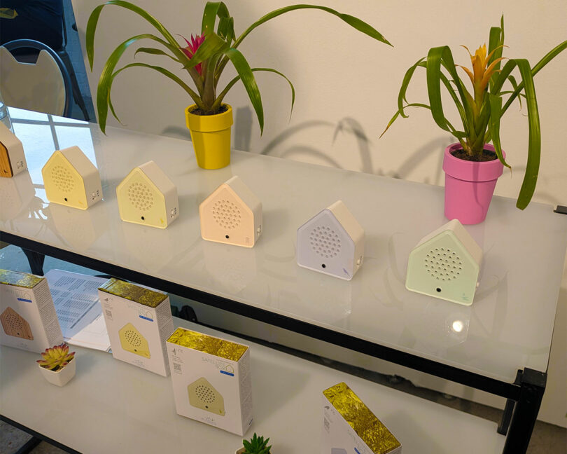 Las exhibiciones en Shoppe Object 2026 presentan filas de pequeños dispositivos electrónicos con forma de casas, plantas en macetas y productos en cajas, todo contra paredes blancas y nítidas.