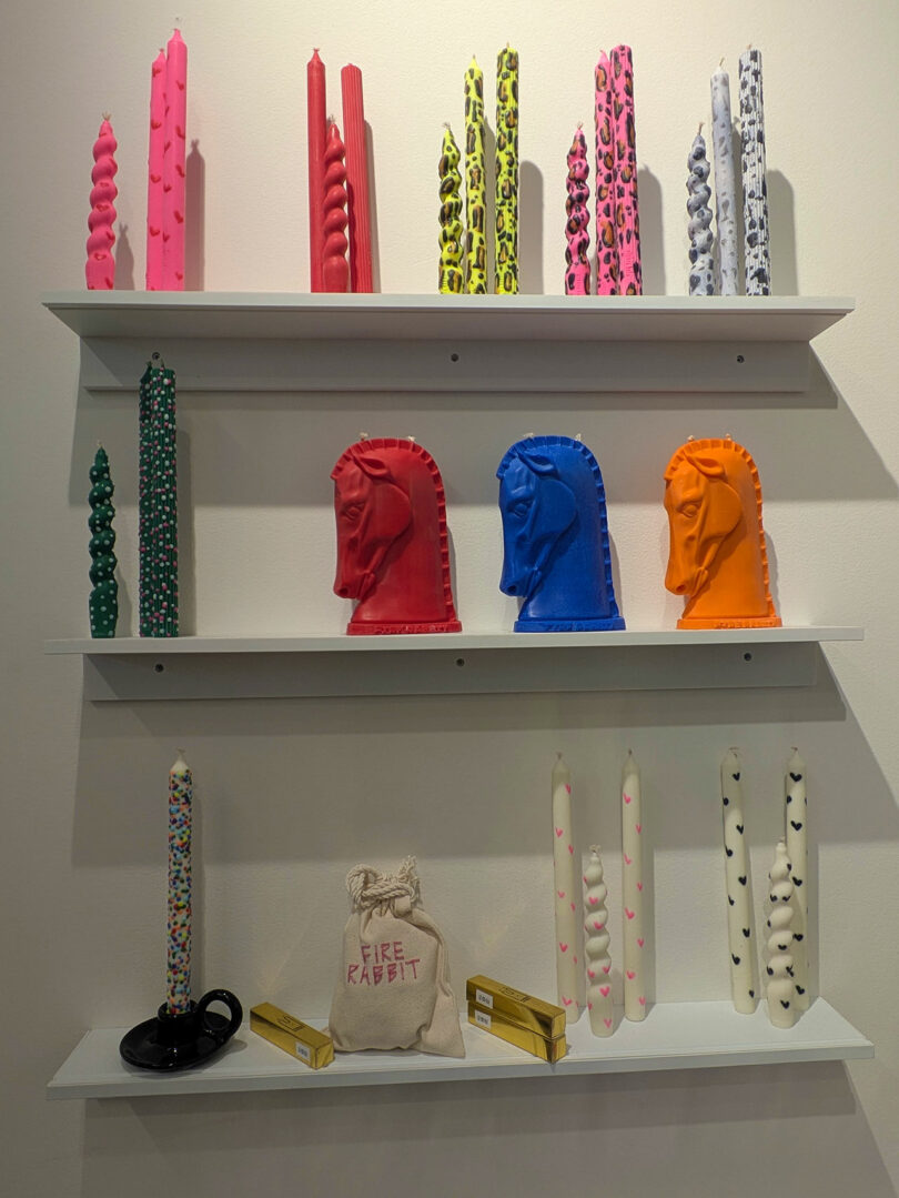 Tres estantes exhiben velas coloridas y con formas únicas, incluidas velas Twist, Pattern y Knight's Head, junto con una pequeña bolsa etiquetada. "conejo de fuego" y dos cajas de cerillas amarillas, todas diseñadas por Shoppe Object 2026.
