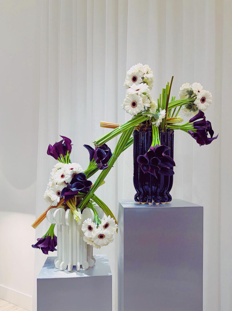 Two modern vases on pedestals holding white gerbera daisies and dark purple calla lilies, arranged in a diagonal, sculptural design against a white curtain backdrop.